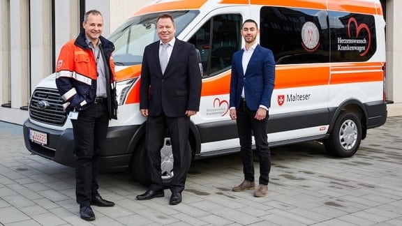 Drei Menschen, davon zwei im Anzug und einer in Rettungsuniform vor einem Rettungswagen 