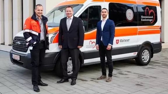 Drei Menschen, davon zwei im Anzug und einer in Rettungsuniform vor einem Rettungswagen 