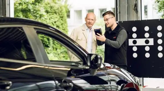 Monteur erklärt Kunde die Kalibrierung eines Fahrzeugs. Monteur hält Gerät in der Hand und zeigt auf das Auto